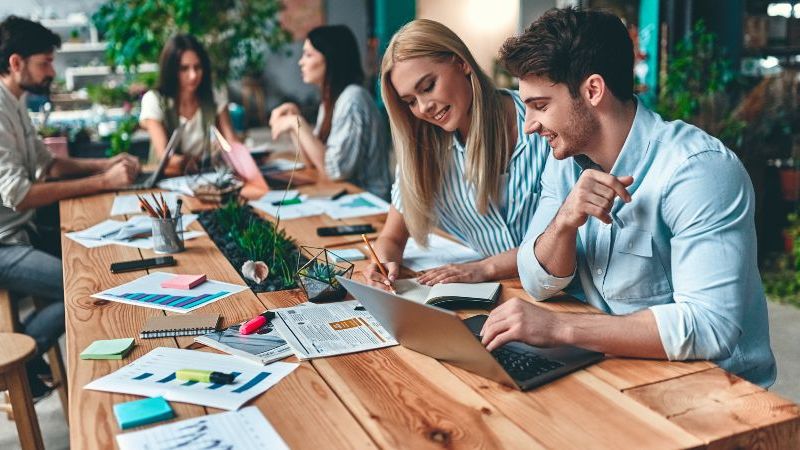 Das Bild zeigt einen Tisch in einem Office-Space, an dem mehre Menschen sitzen und arbeiten.