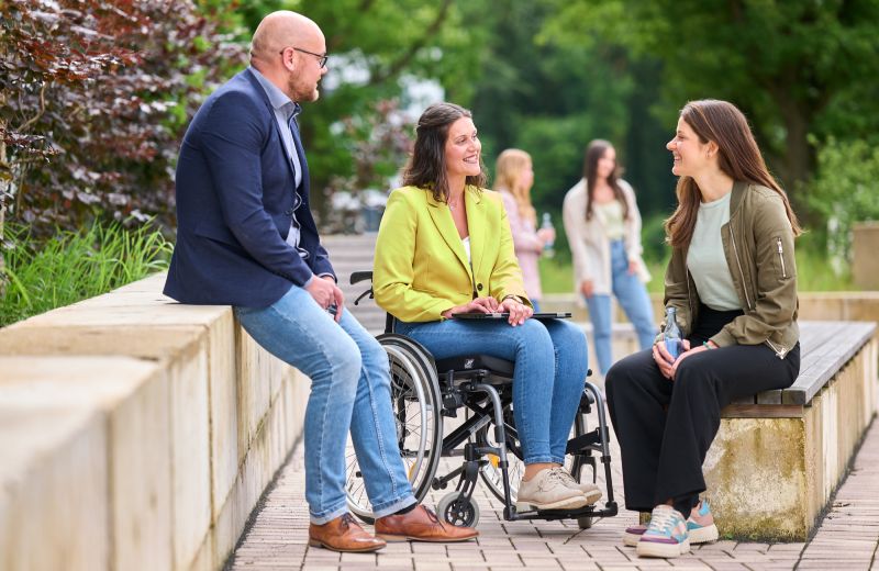 Drei Personen unterhalten sich. Zwei Frauen und ein Mann sitzen auf einer Mauer bzw. einer Bank. Eine Frau sitzt in einem Rollstuhl. Alle wirken fröhlich und entspannt. Im Hintergrund sind noch weitere Personen zu sehen.