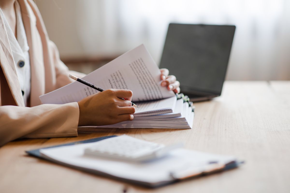 Eine Person sitzt an einem Tisch und schreibt Notizen auf einen Stapel Papier. Im Hintergrund steht ein Laptop.