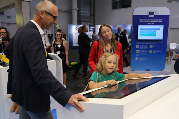 Das Bild zeigt einen Messestand. Im Vordergrund ist ein, in einem weißen Tisch einge-lassener Bildschirm zu sehen, um den sich drei interessiert schauende Personen versammelt haben und mit diesem interagieren. Im Hintergrund sieht man weitere Messestände die von mehreren Menschen umgeben sind. 