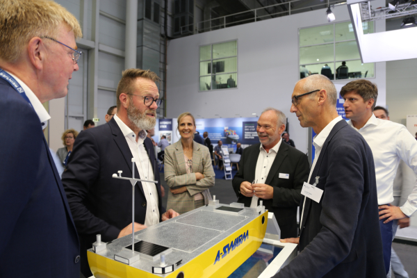 Das Bild zeigt einen Messestand, auf dessen Tisch ein großes gelbes Schiffsmodell ausgestellt wird und von mehreren Menschen, die miteinander reden, umstellt ist. Im Hintergrund sieht man einen weiteren Messestand. 