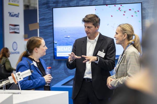 Das Bild zeigt drei Menschen die auf einem Messestand miteinander reden. Im Hintergrund sieht man eine Wand, an der ein großer Bildschirm hängt, sowie ein kleinerer Bildschirm in einem Pult darunter. Im Vordergrund sieht man ein ausgestelltes Objekt aus Metall auf einem Tisch. 