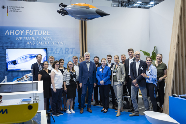 Das Bild zeigt mehrere Menschen, die vor einem Messestand für ein Gruppenfoto zusammenstehen. Von der Decke herunter hängt ein Modell in Form eines kleinen Unterwasserfahrzeugs. 