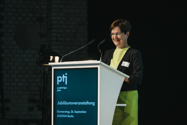 Das Bild zeigt PtJ-Leiterin Dr. Stephanie Bauer hinter einem Podium mit Mikrofonen und der Aufschrift „Jubiläumsveranstaltung“. 